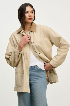 A woman models the ROSA JACKET by Velvet by Graham & Spencer—a beige cotton canvas jacket styled over a white top and blue jeans, with one hand in her pocket against a plain background, ideal for effortless everyday looks.