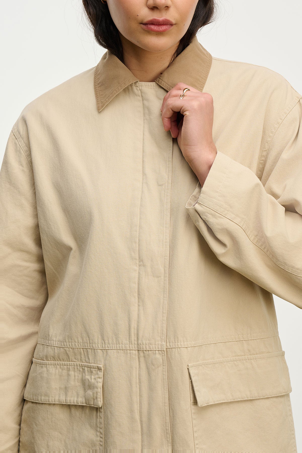 A person wearing the oversized beige ROSA JACKET by Velvet by Graham & Spencer, featuring large front pockets and a relaxed fit. Their face is partially visible as they hold the collar, showcasing this stylish cotton canvas piece.-39654938509505