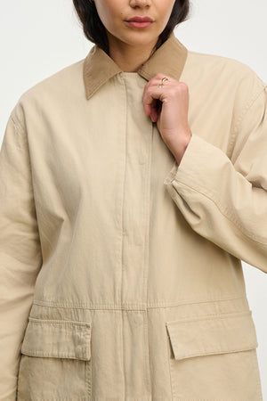 A person wearing the oversized beige ROSA JACKET by Velvet by Graham & Spencer, featuring large front pockets and a relaxed fit. Their face is partially visible as they hold the collar, showcasing this stylish cotton canvas piece.