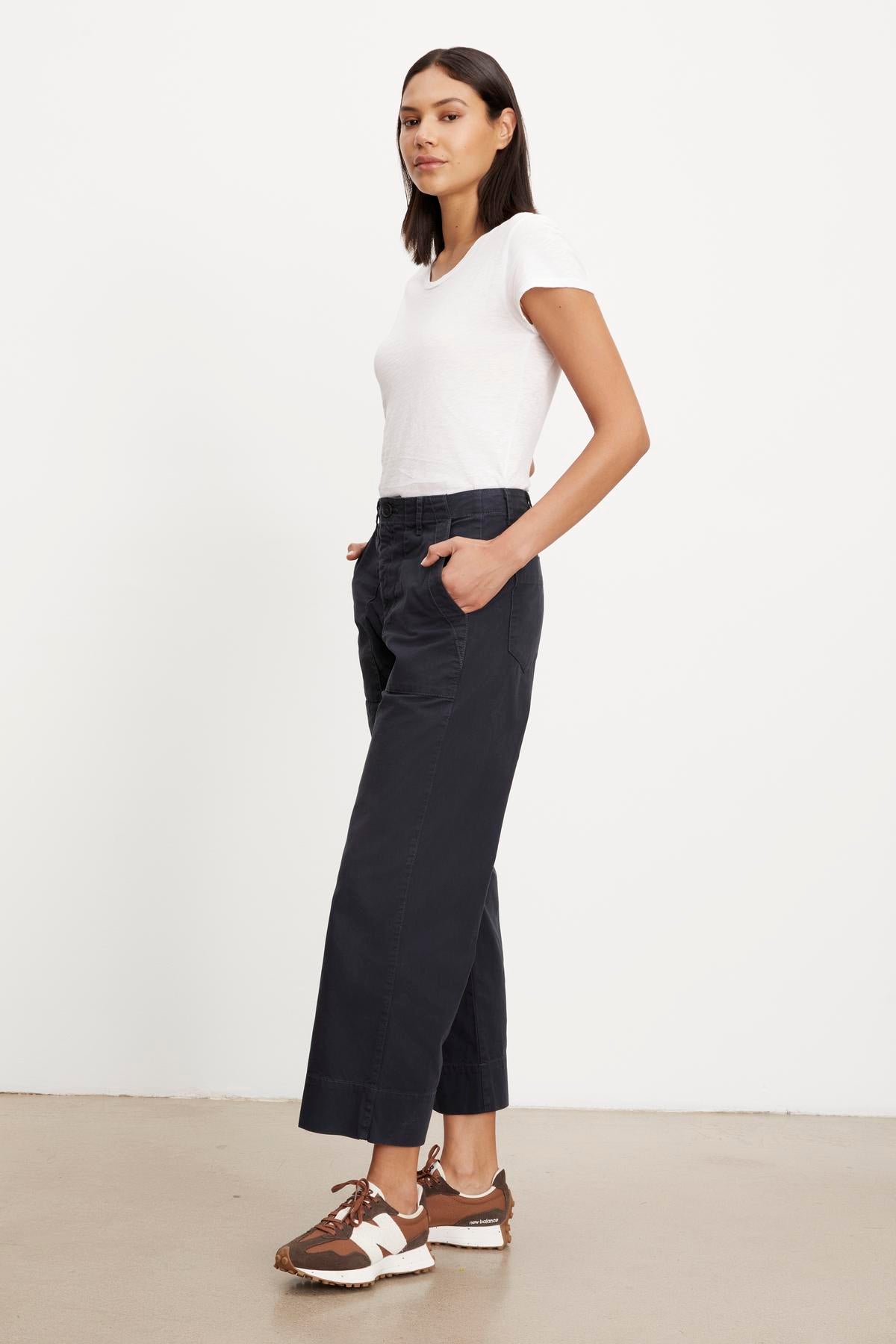   A woman stands casually in a white top, Velvet by Graham & Spencer's MYA cotton canvas pants with a utility silhouette, and brown shoes against a neutral background. 