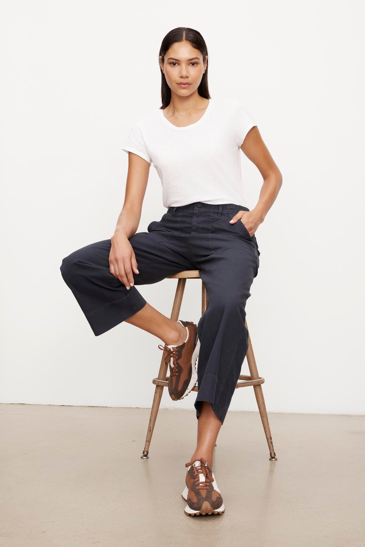   A woman seated on a wooden stool, wearing a white t-shirt and navy garment dye culottes with brown strappy sandals.(Product Name: Velvet by Graham & Spencer MYA COTTON CANVAS PANT.) 