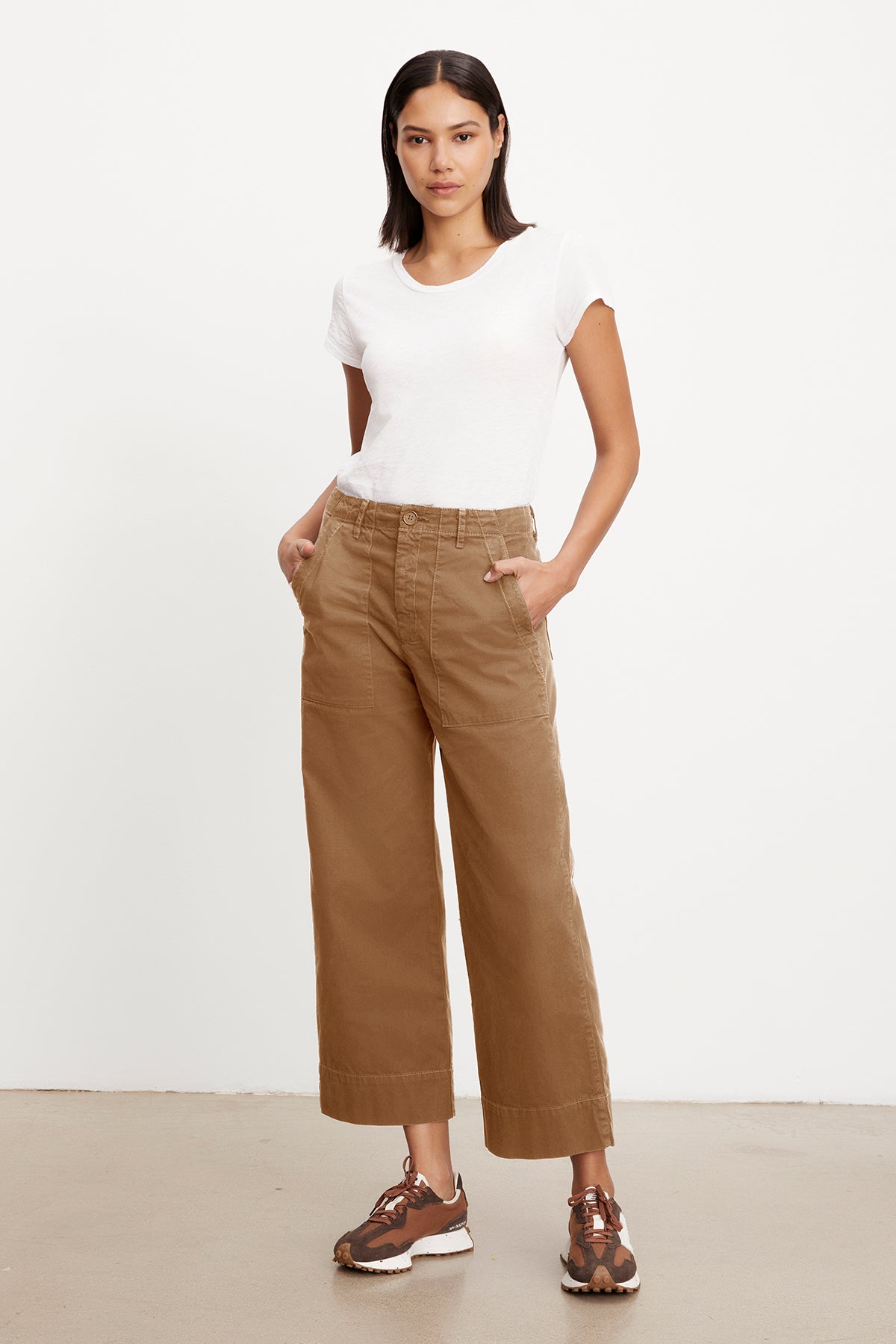   A woman stands against a plain white background, wearing a slim top, MYA COTTON CANVAS PANT by Velvet by Graham & Spencer, and brown sneakers. Her hands are in her pockets, and she gazes directly at the camera. 