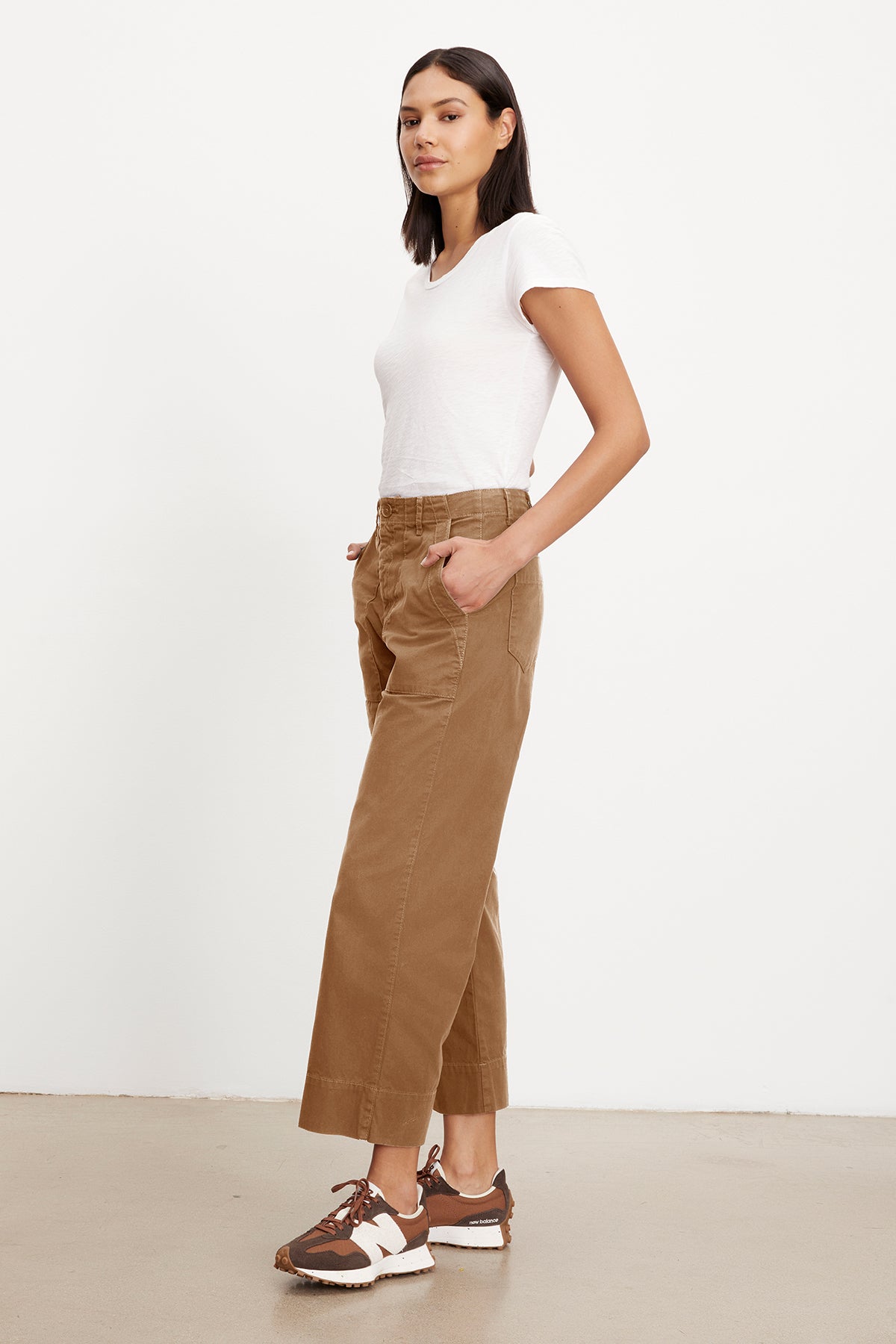 A person stands sideways, hands in pockets, wearing a white slim top, Velvet by Graham & Spencer MYA COTTON CANVAS PANT with a wide-leg cut, and brown sneakers, against a plain white background.-37355950342337