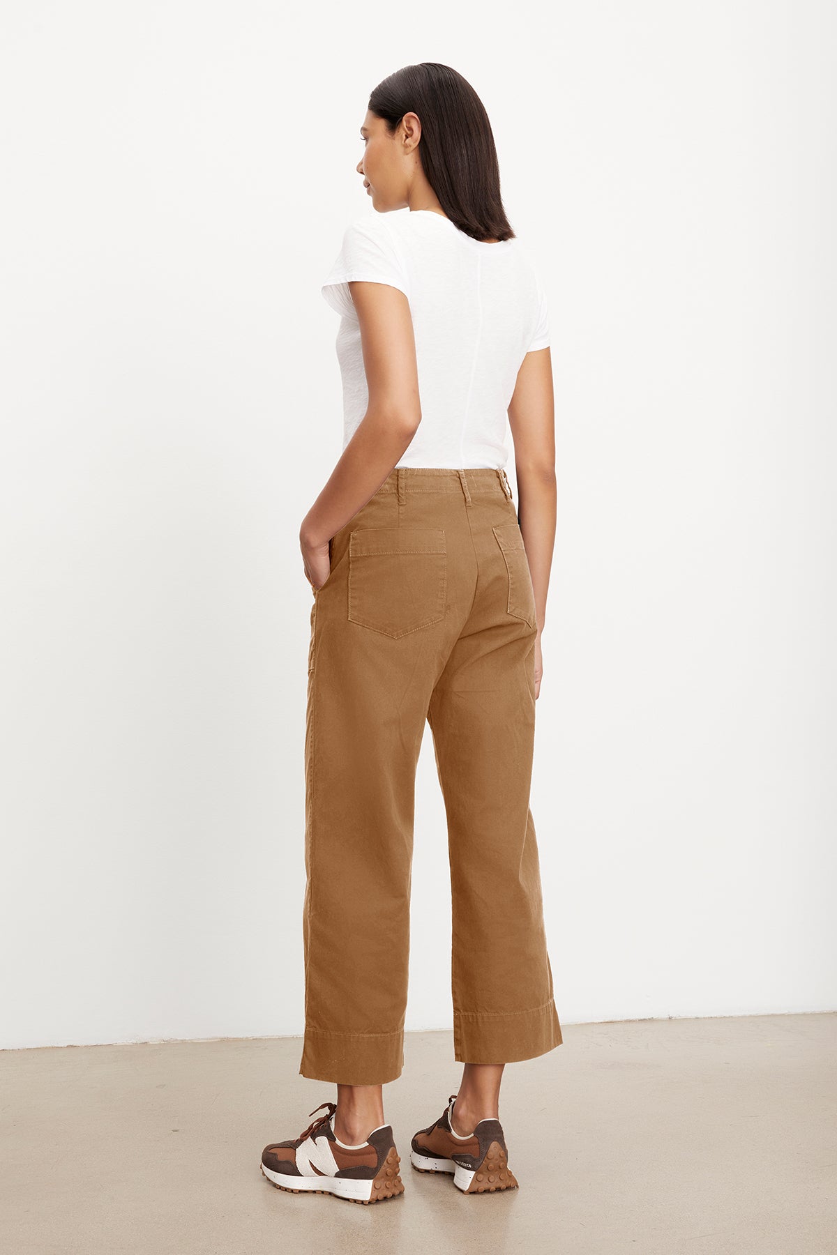   A woman stands facing away, wearing a slim top, MYA COTTON CANVAS PANT by Velvet by Graham & Spencer with a flared leg, and brown sneakers. Her hands are in her pockets as she looks to the right side. 