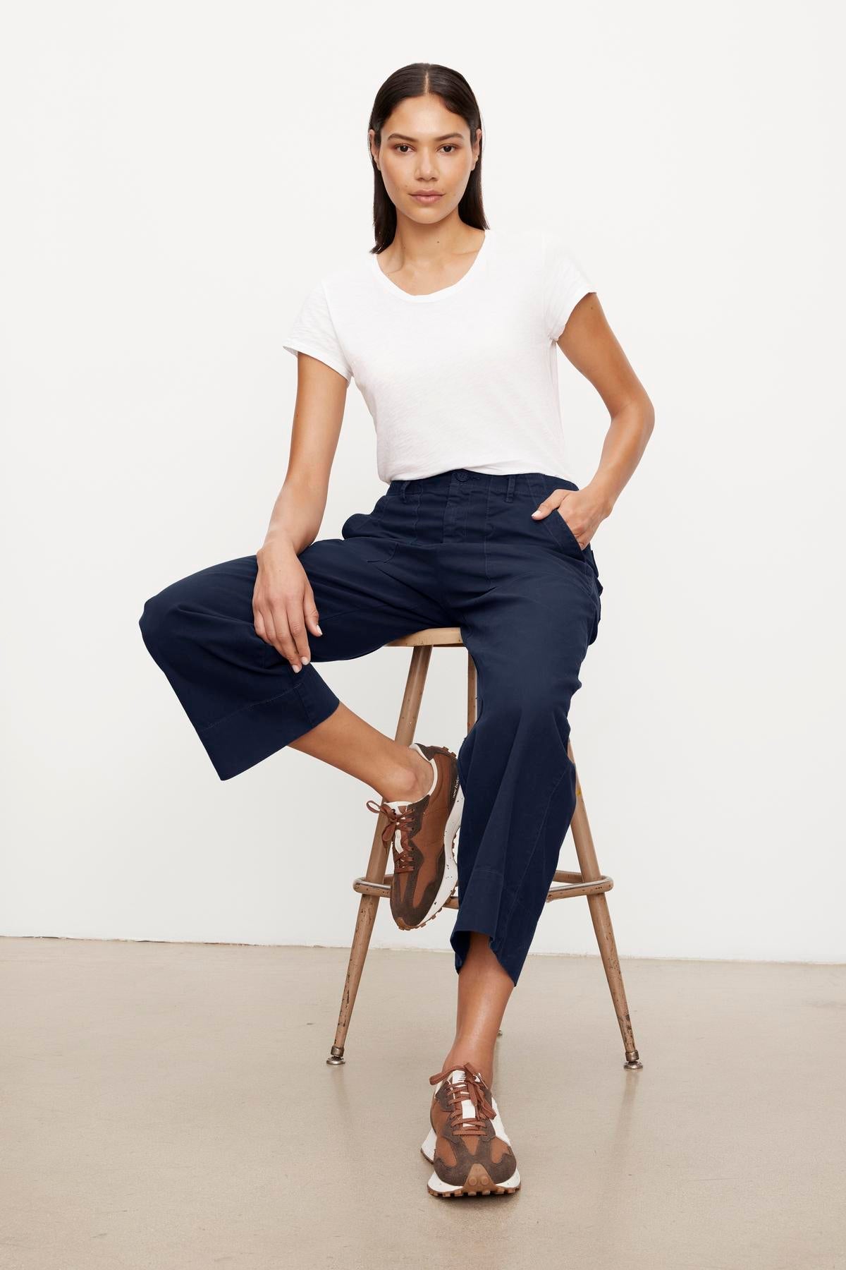 A woman sits on a wooden stool, wearing a white T-shirt and navy blue garment-dyed Velvet by Graham & Spencer culottes with brown sandals, facing the camera in a studio setting.-37628822388929