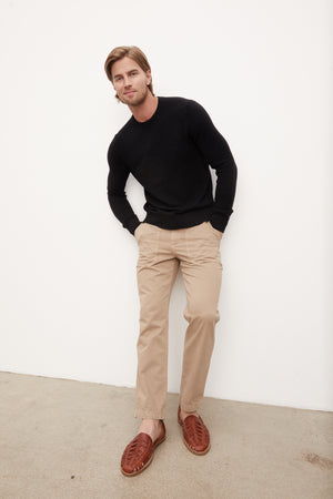 A man in a black Velvet by Graham & Spencer ACE Thermal Crew sweater and tan pants leaning against a wall.