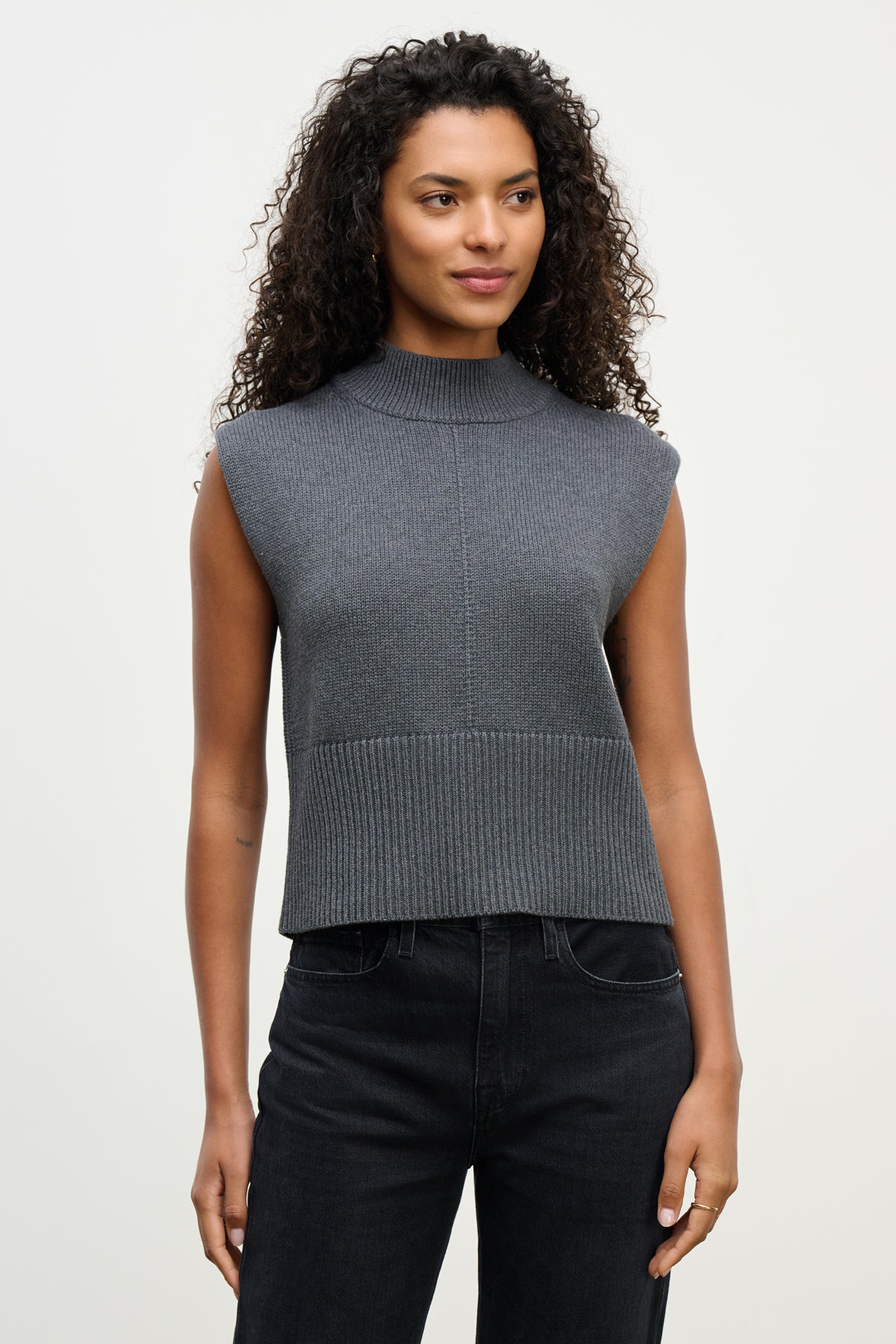 A woman with curly hair wears the ELECTRA SWEATER VEST by Velvet by Graham & Spencer and black jeans, standing against a plain light background.