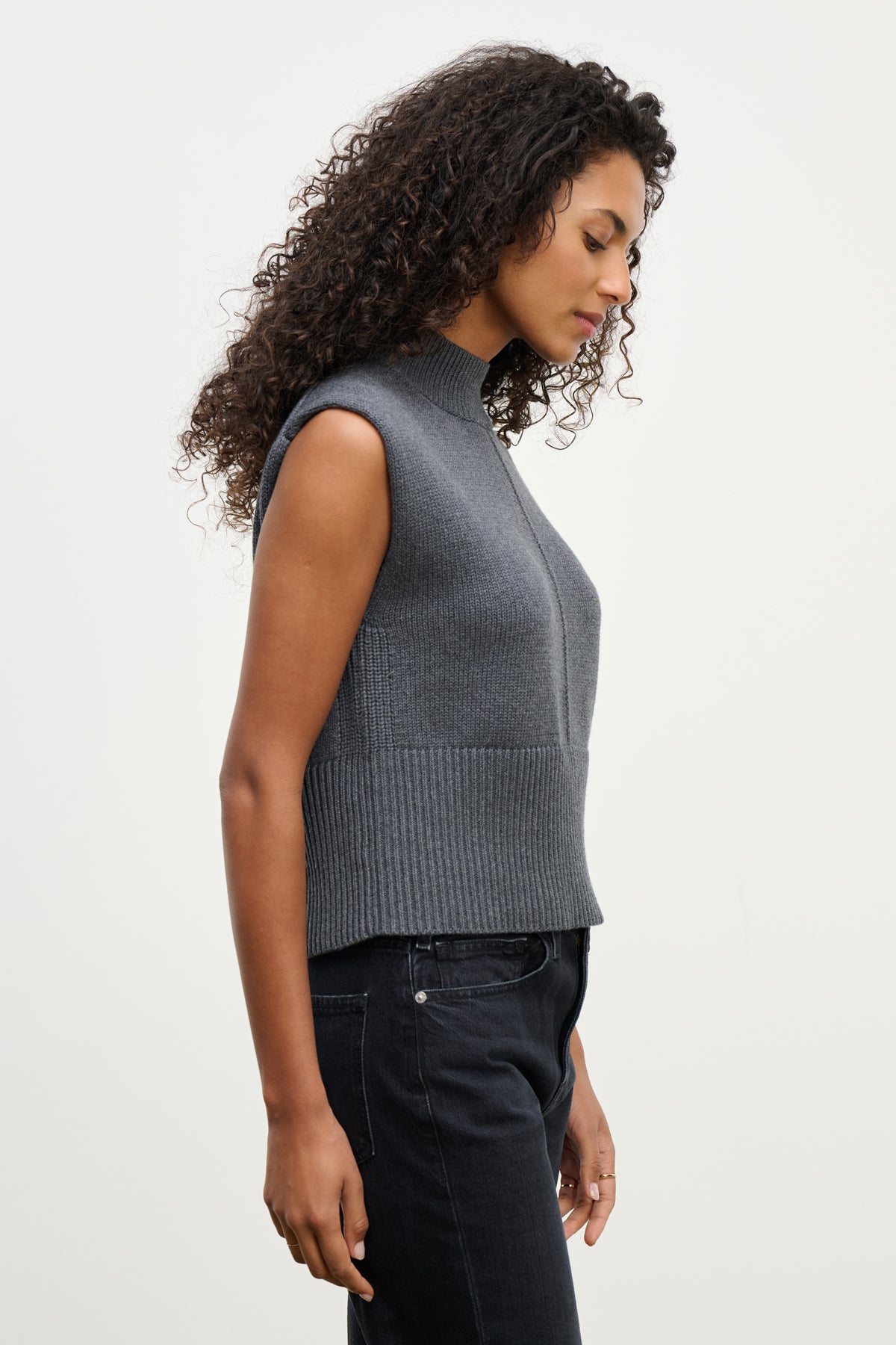 Woman with curly hair models the ELECTRA SWEATER VEST by Velvet by Graham & Spencer—a cotton-cashmere blend mock neck vest with ribbed hems—paired with black pants, shown in profile against a plain light background.-40227049537729