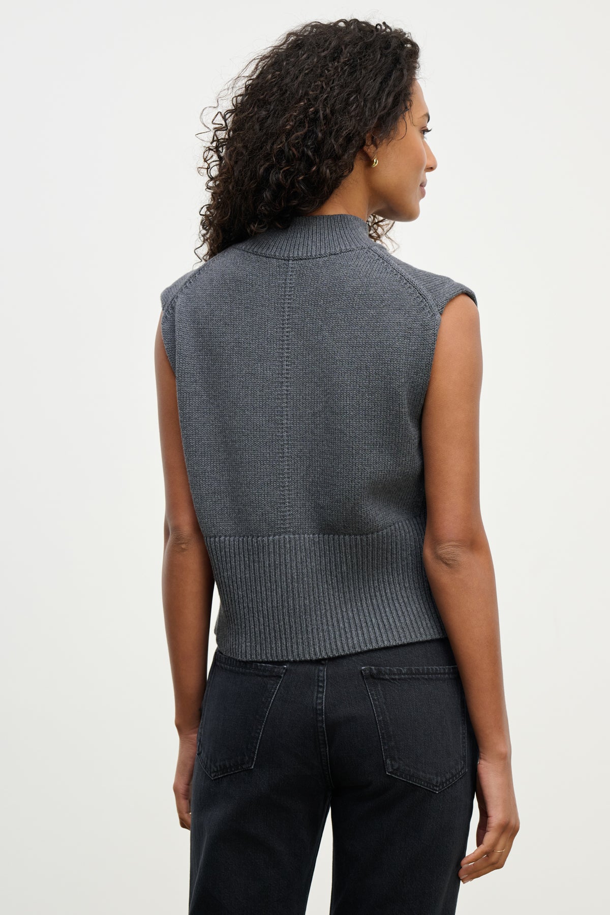 A woman with curly hair is seen from the back wearing the ELECTRA SWEATER VEST by Velvet by Graham & Spencer—a cotton cashmere blend, sleeveless knit with ribbed banded hems—paired with black jeans against a plain light background.-40227049603265