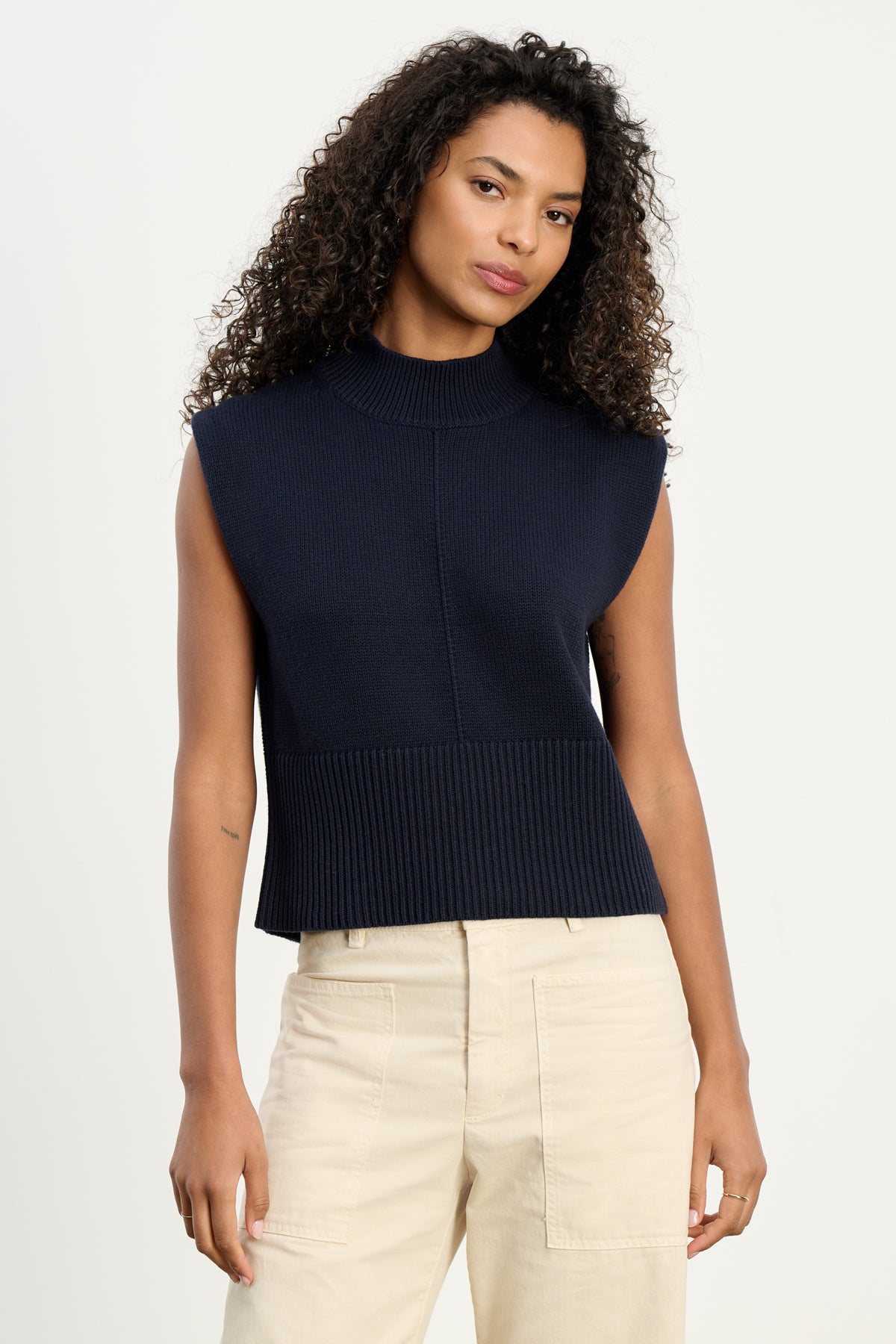   A woman with curly hair wears the ELECTRA COTTON CASHMERE SWEATER VEST by Velvet by Graham & Spencer in navy, styled sleeveless with ribbed hems, paired with light beige pants against a plain white background. 