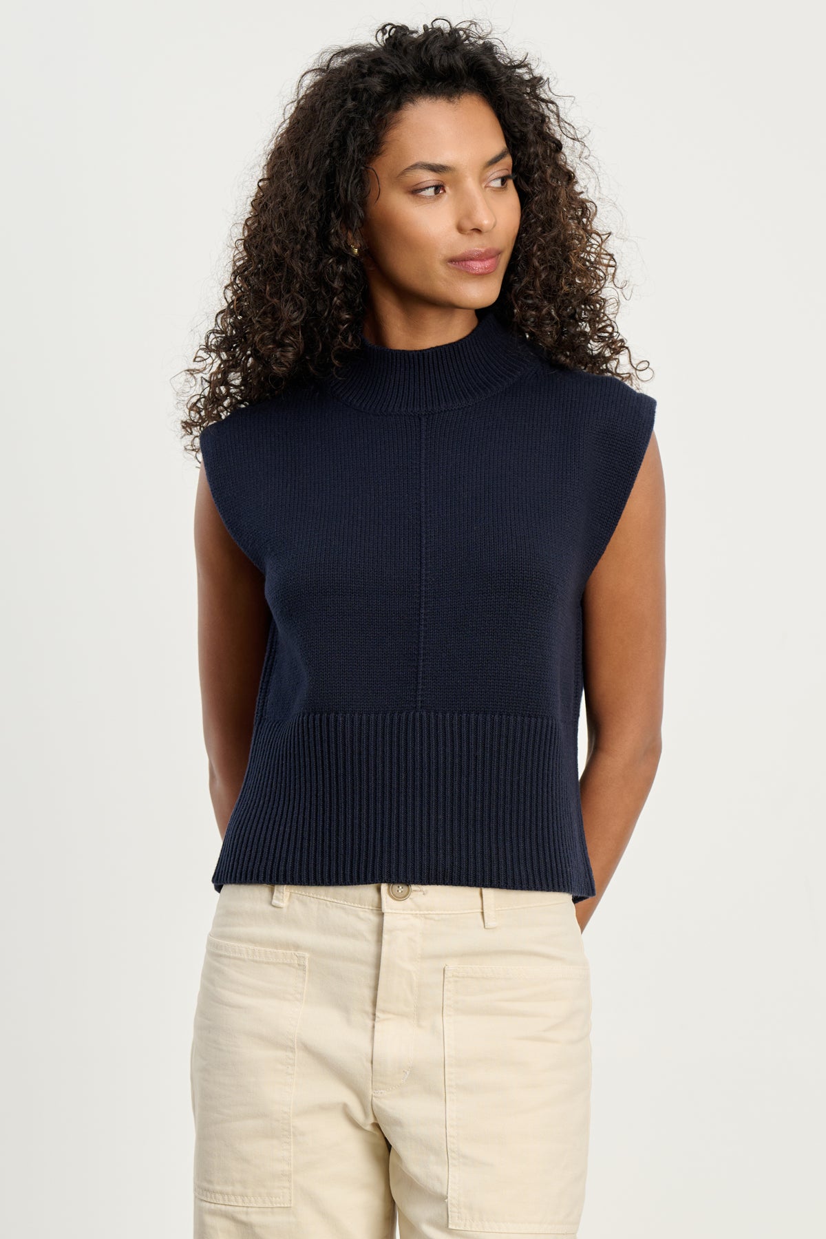   A woman with curly hair wears the ELECTRA COTTON CASHMERE SWEATER VEST by Velvet by Graham & Spencer, featuring ribbed hems, styled with light beige pants against a plain light background. 