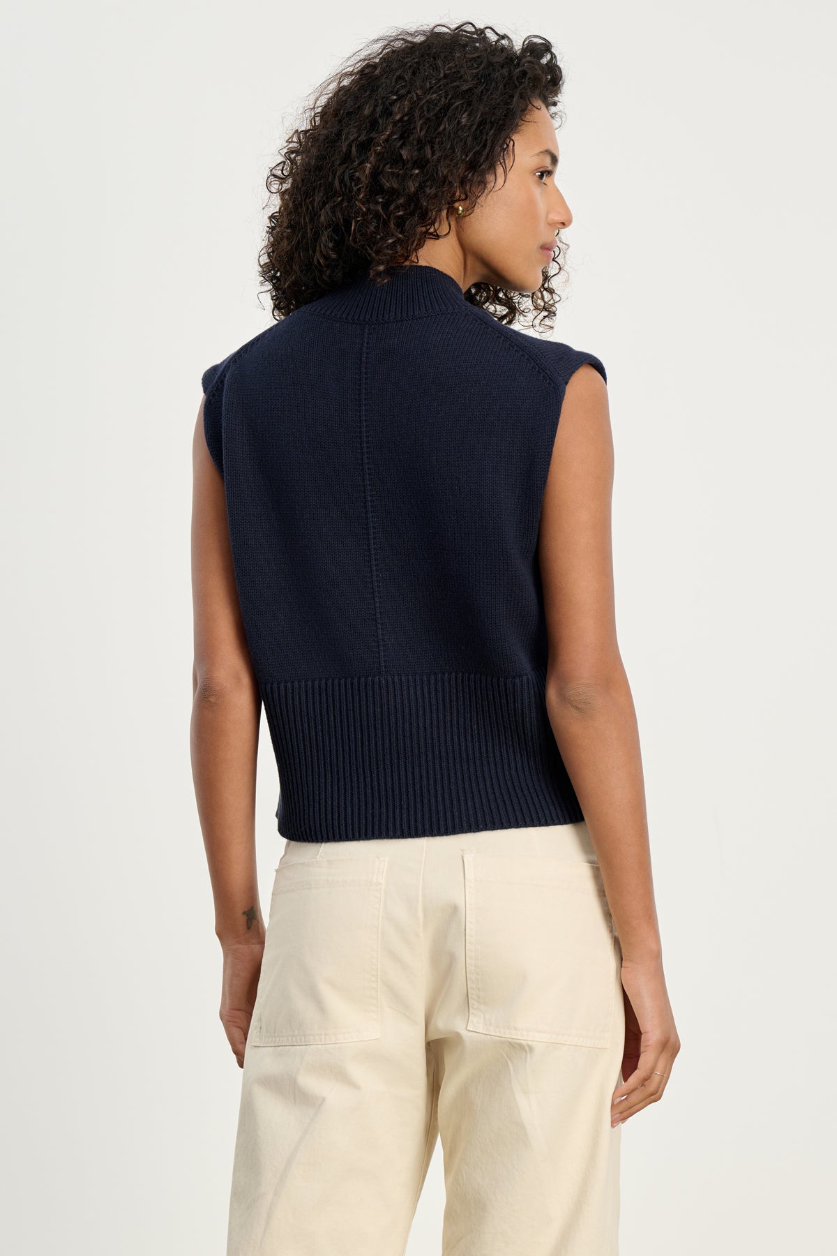 A person with curly hair, dressed in the Velvet by Graham & Spencer ELECTRA COTTON CASHMERE SWEATER VEST in navy and light beige pants, stands facing away from the camera against a plain background.-39800299323585