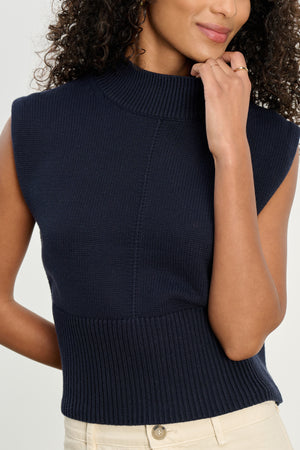 A woman in the Velvet by Graham & Spencer ELECTRA Cotton Cashmere Sweater Vest, a sleeveless navy knit turtleneck with ribbed hems, pairs it with beige pants and stands with one hand near her chin.