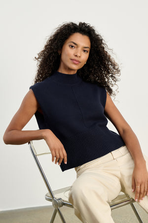 A person with curly hair wears the ELECTRA COTTON CASHMERE SWEATER VEST by Velvet by Graham & Spencer, styled sleeveless in navy, paired with cream pants while seated on a metal chair against a plain background.