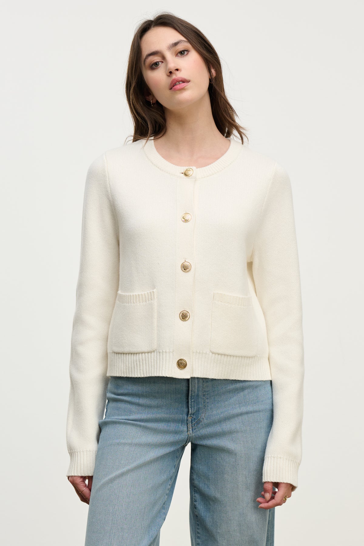 A woman wears the MAHAL CARDIGAN by Velvet by Graham & Spencer, featuring embellished buttons and front pockets, paired with light blue jeans, against a plain light background.-40227113369793