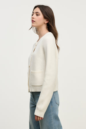 A woman stands in profile against a white background, modeling the MAHAL CARDIGAN by Velvet by Graham & Spencer—a versatile cotton-cashmere piece with embellished buttons—paired with blue jeans.
