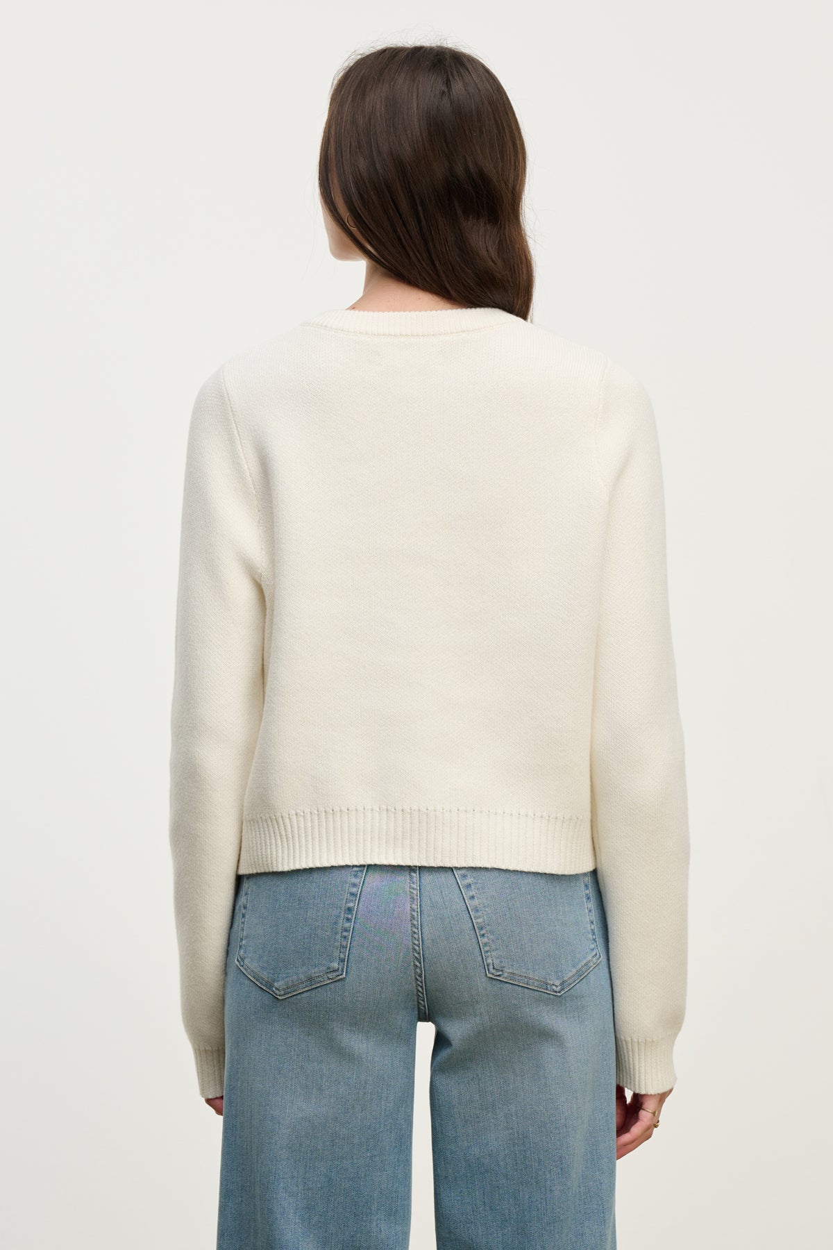 A person with long brown hair wears the MAHAL CARDIGAN by Velvet by Graham & Spencer—an embellished cream cotton-cashmere layer—paired with light blue jeans, shown from the back against a plain background.-40227113435329