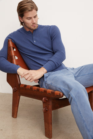 A man sitting on a wooden chair wearing a blue Velvet by Graham & Spencer JAKE THERMAL HENLEY sweater.