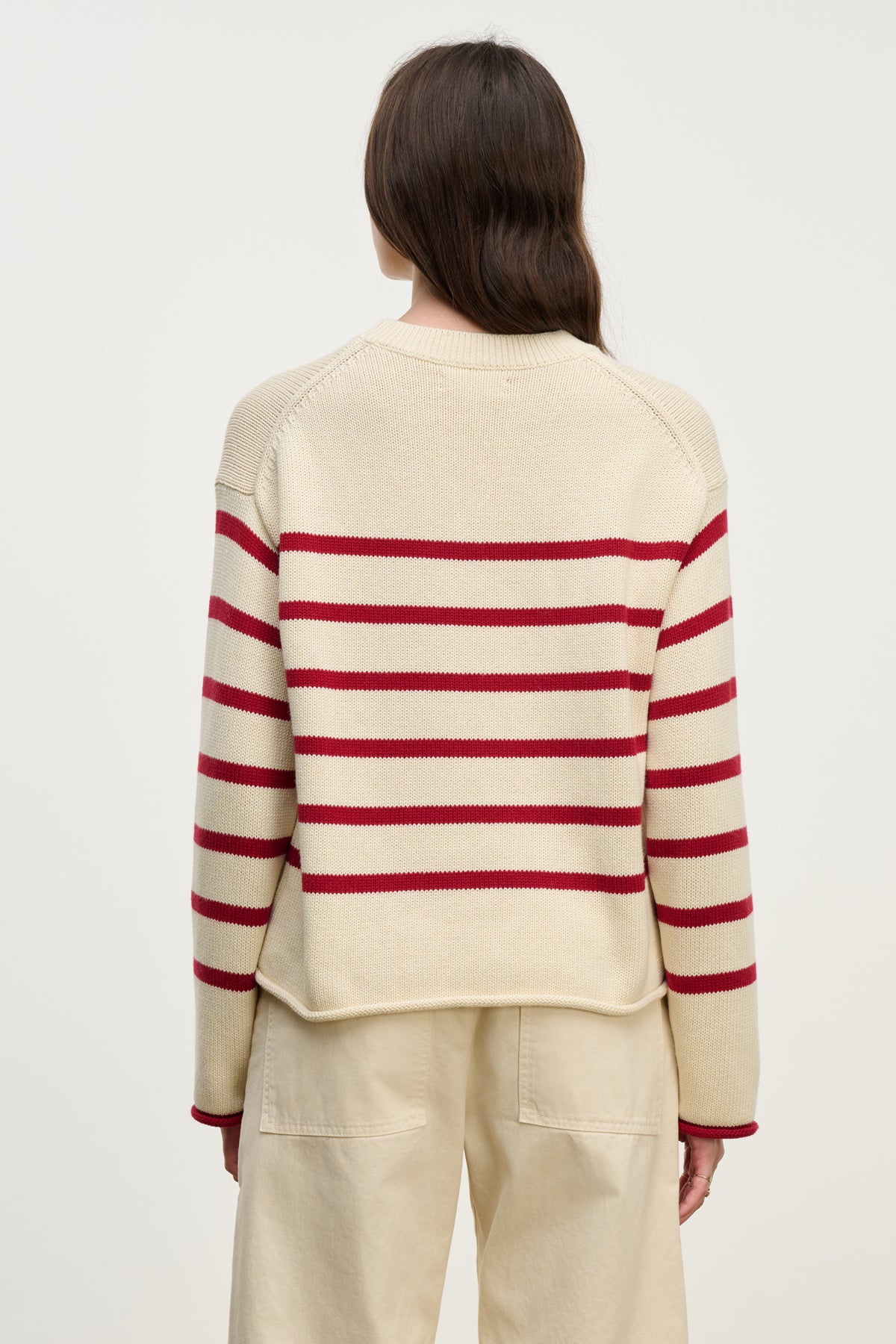 A person stands with their back to the camera, wearing the LUCIA SWEATER by Velvet by Graham & Spencer—this relaxed-fit cotton-cashmere sweater features horizontal red stripes and a classic crew neckline, paired with cream pants against a plain light background.