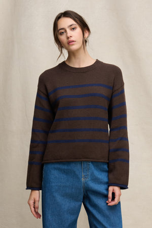A person wears the LUCIA COTTON CASHMERE SWEATER by Velvet by Graham & Spencer—a brown, relaxed-fit crewneck with navy stripes—paired with blue jeans, standing before a beige fabric backdrop.