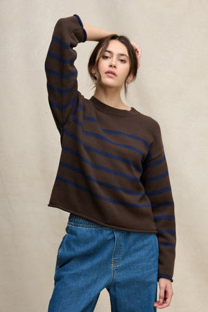 A person stands against a neutral background wearing the LUCIA COTTON CASHMERE SWEATER by Velvet by Graham & Spencer, featuring blue stripes and a relaxed fit, paired with blue jeans and raising one hand to their head.