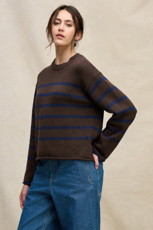 A woman wears the LUCIA COTTON CASHMERE SWEATER by Velvet by Graham & Spencer— a relaxed-fit brown crewneck with blue stripes—paired with blue jeans, standing before a beige background.