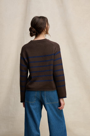 A person stands facing away, wearing the Velvet by Graham & Spencer LUCIA Cotton Cashmere Sweater with blue horizontal stripes and blue jeans, set against a neutral backdrop.
