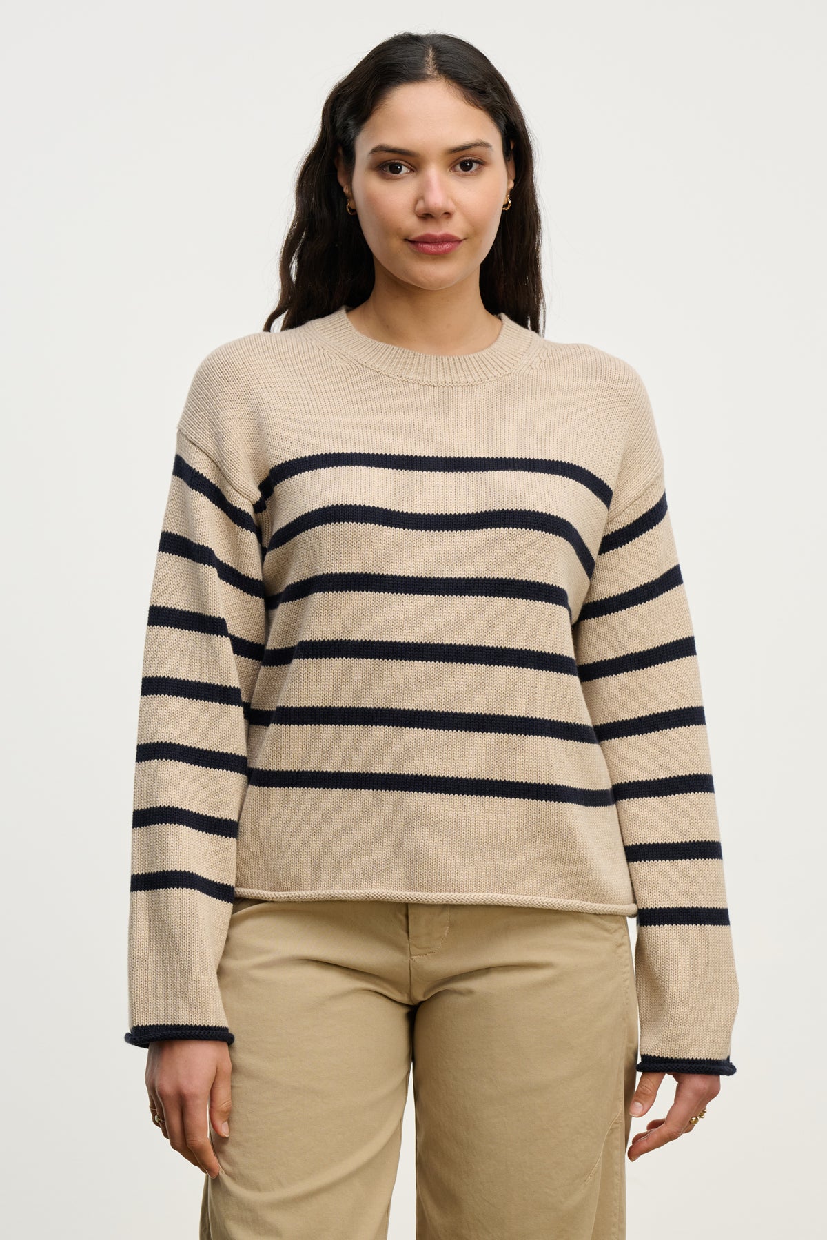A woman stands against a plain background wearing the LUCIA SWEATER by Velvet by Graham & Spencer—a beige, relaxed-fit crewneck with navy stripes—paired with beige pants.-40227097706689