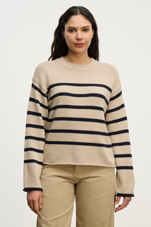 A woman stands against a plain background wearing the LUCIA SWEATER by Velvet by Graham & Spencer—a beige, relaxed-fit crewneck with navy stripes—paired with beige pants.