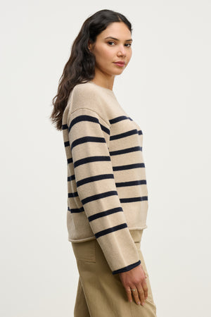 A woman with long dark hair stands sideways against a plain light background wearing the LUCIA SWEATER by Velvet by Graham & Spencer—a relaxed-fit beige cotton-cashmere sweater with navy stripes—paired with khaki pants.