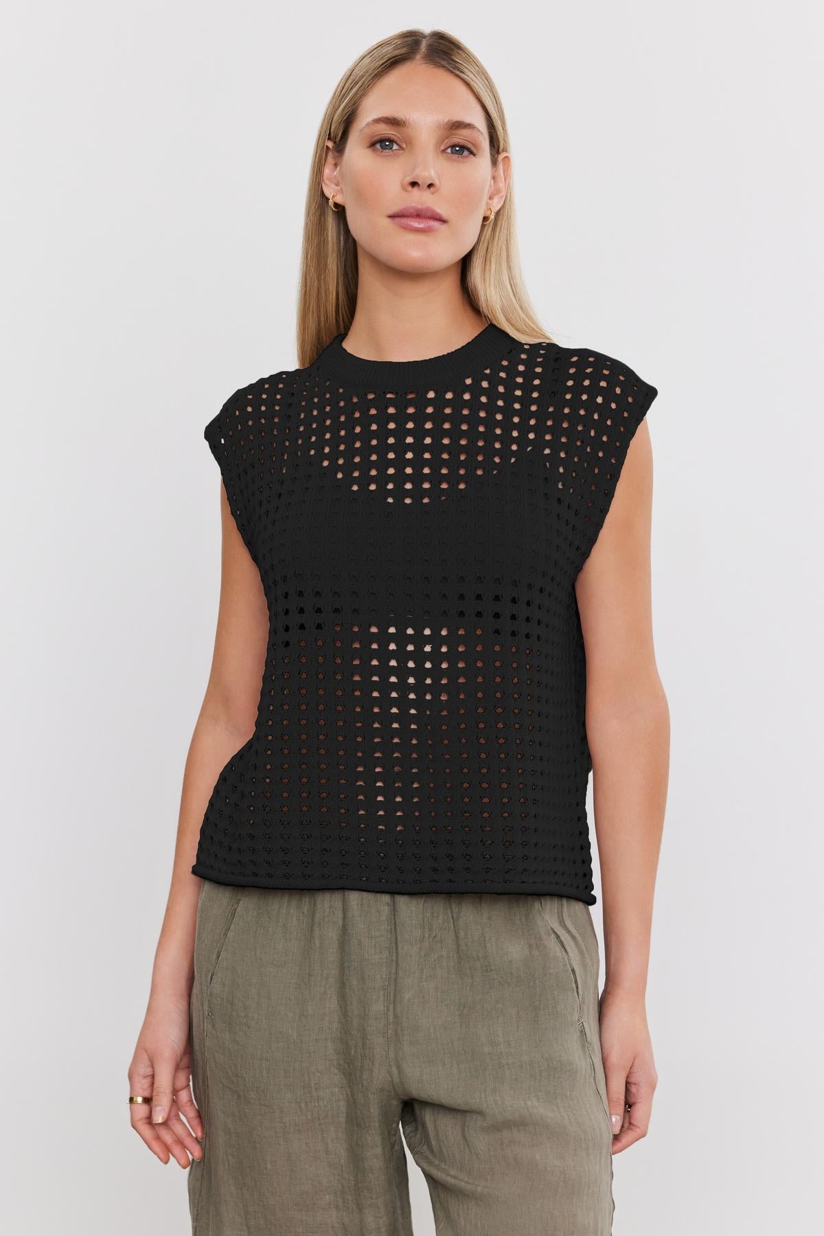  A person with long blonde hair is wearing a black, sleeveless, eyelet knit MAISON SWEATER by Velvet by Graham & Spencer over a black bra, paired with loose-fitting khaki pants. The top features a cropped silhouette perfect for summer. The background is plain white. 