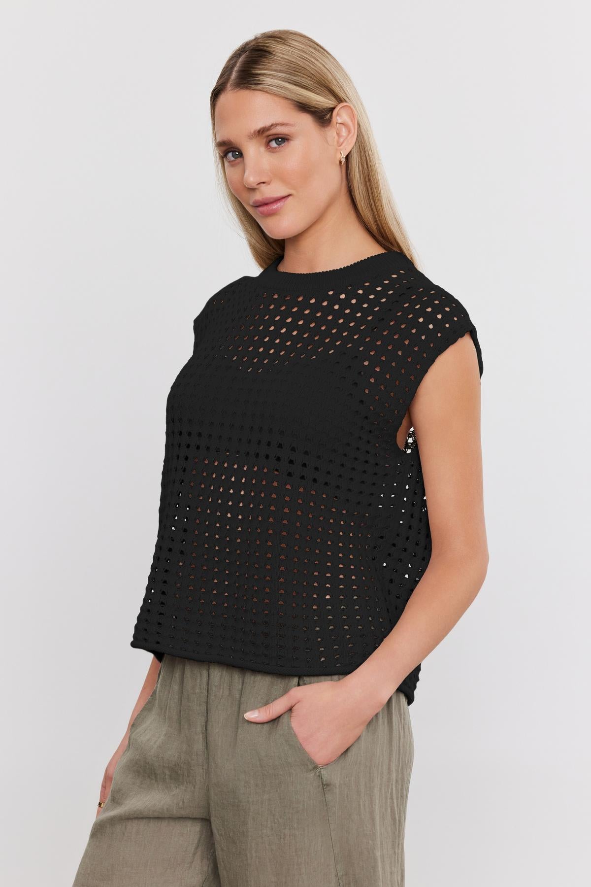 A person with long blonde hair wearing a black perforated top and khaki pants stands against a plain white background, showcasing the stylish MAISON SWEATER by Velvet by Graham & Spencer.-37054537892033