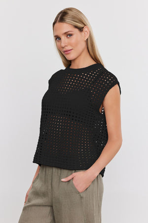 A person with long blonde hair wearing a black perforated top and khaki pants stands against a plain white background, showcasing the stylish MAISON SWEATER by Velvet by Graham & Spencer.