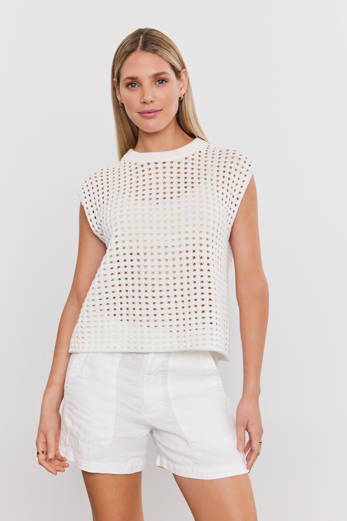  A woman with long blonde hair, wearing the MAISON SWEATER by Velvet by Graham & Spencer and white shorts, stands against a plain white background, showcasing her chic cropped silhouette. 