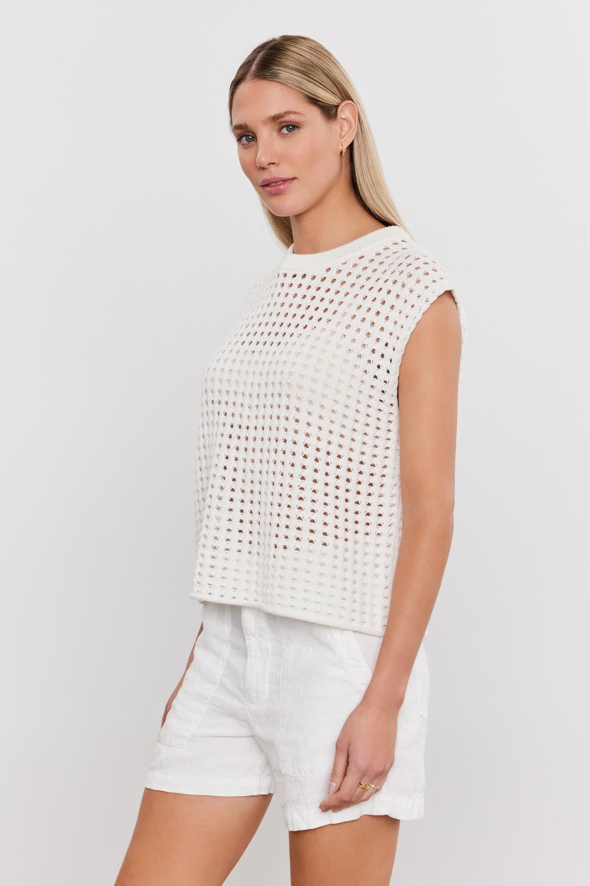   A person with long blond hair is wearing a white sleeveless MAISON SWEATER by Velvet by Graham & Spencer and white shorts, standing against a plain white background. 