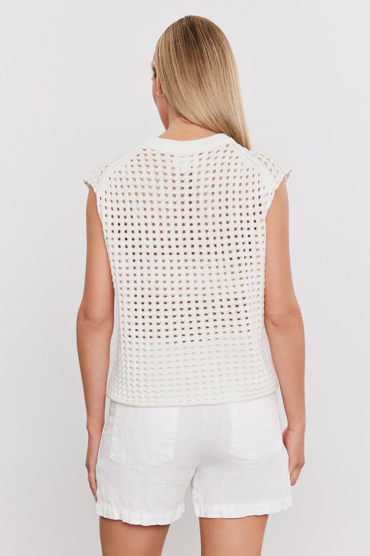   Person with long blond hair, wearing a white, sleeveless, crocheted top and white shorts, standing with their back facing the camera against a plain white background, presenting a cropped silhouette ideal for showcasing Velvet by Graham & Spencer's MAISON SWEATER in lightweight cotton cashmere. 