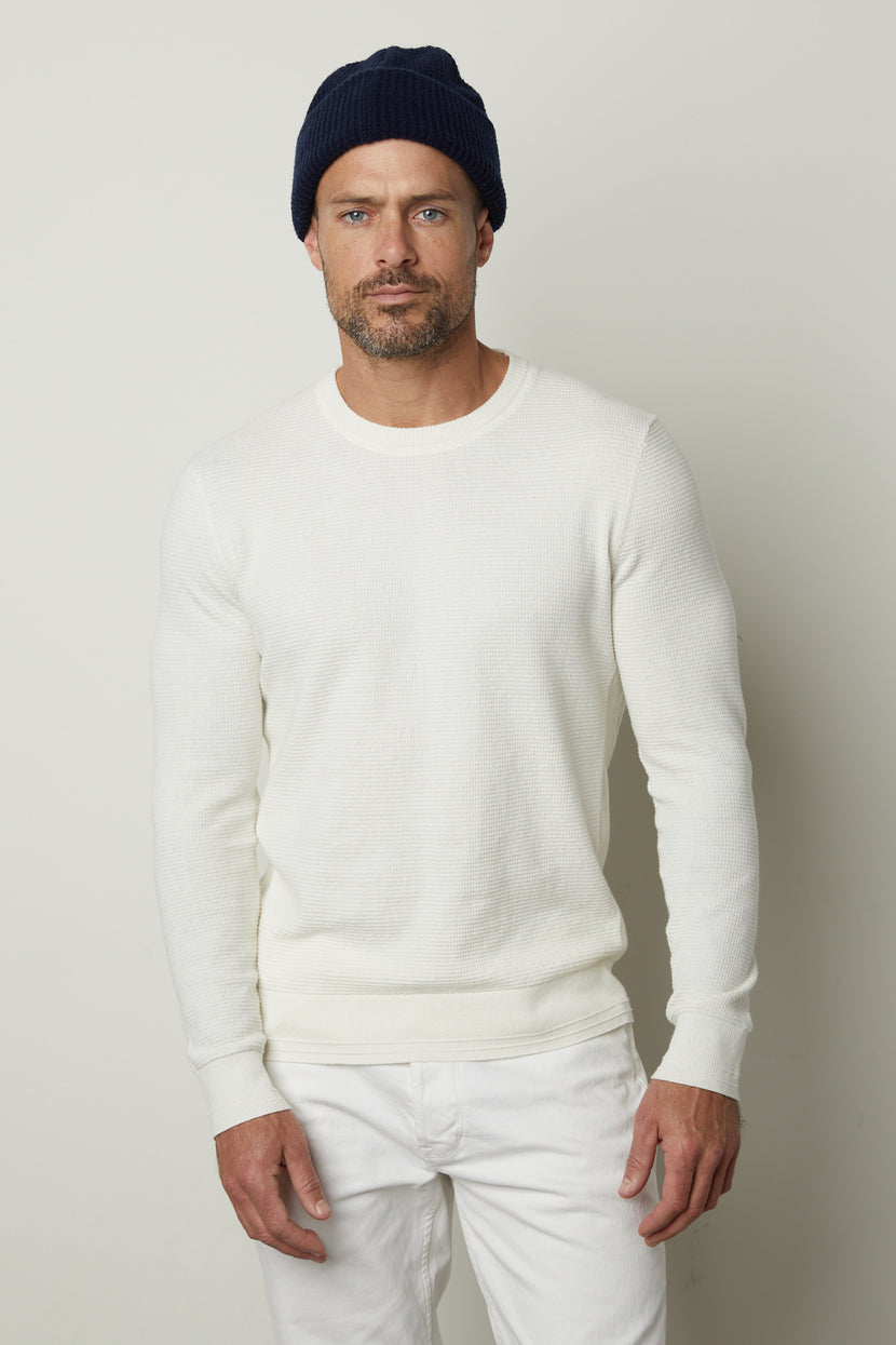 A man stands facing the camera wearing a navy blue beanie, a white WALTER SWEATER by Velvet by Graham & Spencer featuring a thermal design, and white pants.