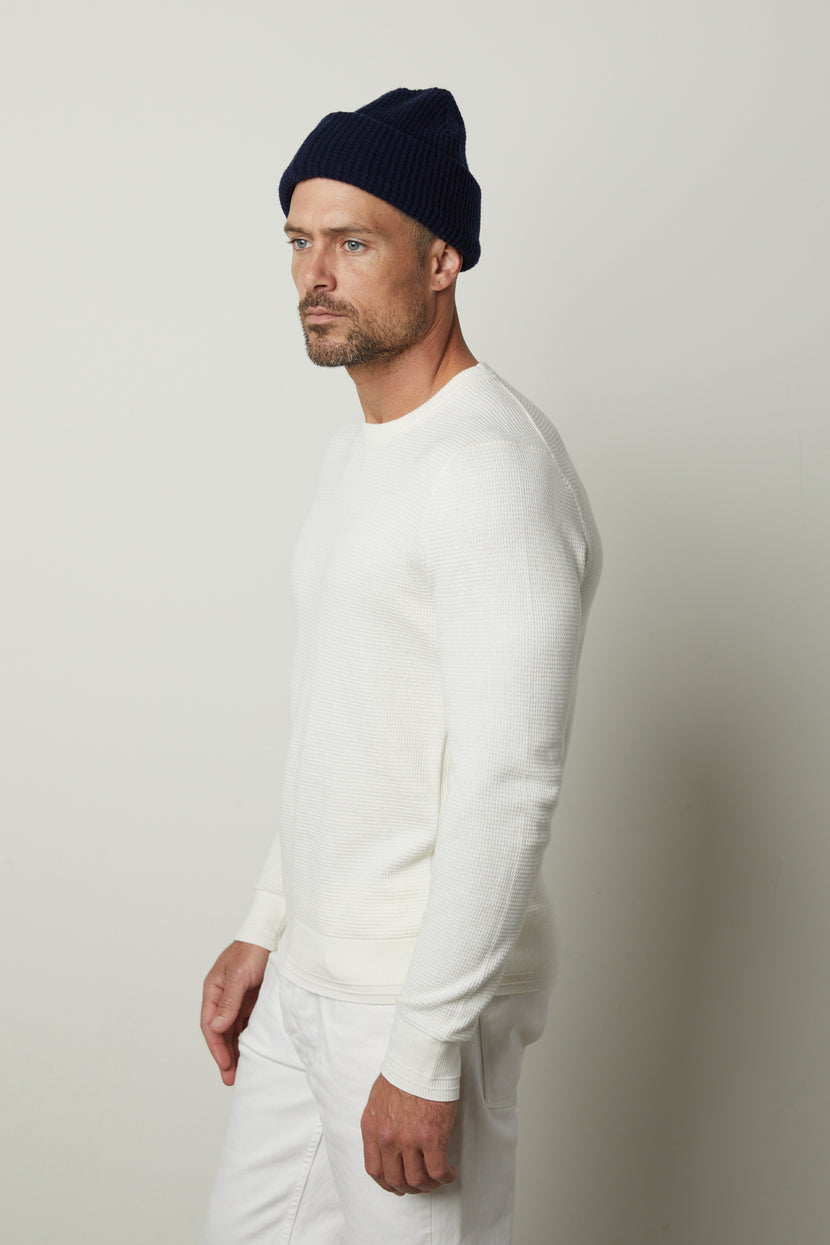 A man with a beard, wearing a navy knit beanie and the WALTER SWEATER—a white long-sleeve shirt made of soft cotton by Velvet by Graham & Spencer—paired with white pants, stands against a plain white background, looking to the left.