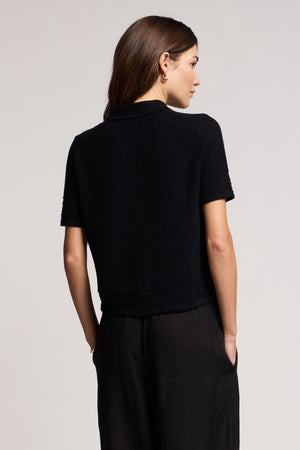 A woman with long brown hair, seen from behind, wears the Velvet by Graham & Spencer ROSEMOND CARDIGAN—a short-sleeved black knit with mesh stitching—and black pants, hands in her pockets, against a plain light background.
