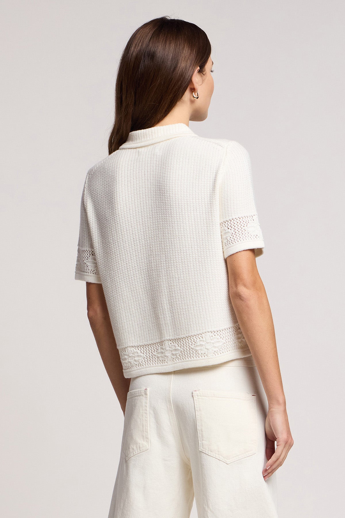 A woman with long brown hair stands facing away from the camera, wearing Velvet by Graham & Spencer's ROSEMOND CARDIGAN—a short-sleeve white knit top with mesh stitching—paired with white pants.-41372113076417