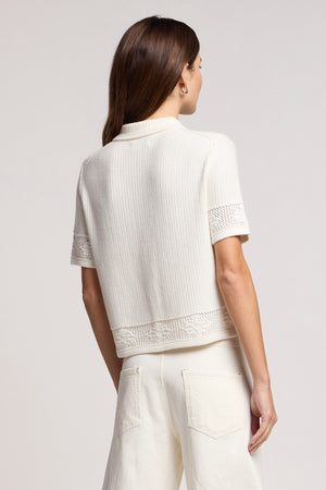 A woman with long brown hair stands facing away from the camera, wearing Velvet by Graham & Spencer's ROSEMOND CARDIGAN—a short-sleeve white knit top with mesh stitching—paired with white pants.