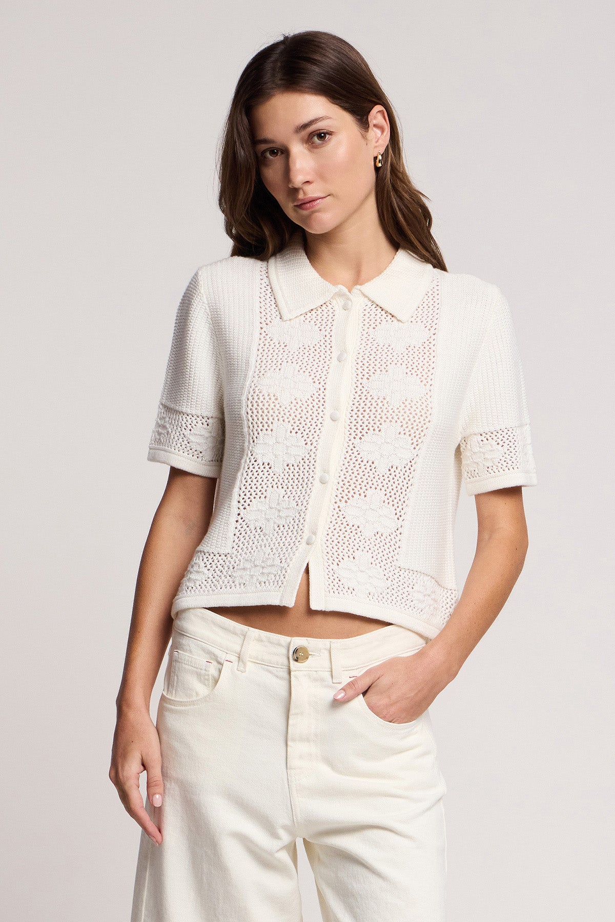 A woman wears the ROSEMOND CARDIGAN by Velvet by Graham & Spencer—a short-sleeve, button-up cream crochet top with a collar—paired with high-waisted cream pants, standing against a plain background.-41372113109185
