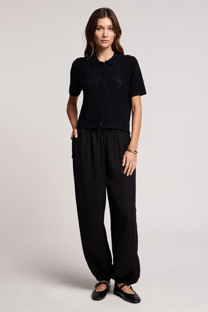 A person stands facing forward, hands in pockets, wearing a short-sleeve black shirt, Velvet by Graham & Spencer CAMBER LINEN PANT with an elastic waist, and black flat shoes against a plain background.