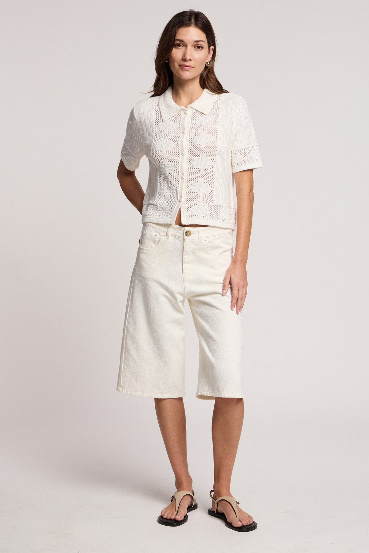 A woman faces forward against a plain background, wearing the ROSEMOND CARDIGAN by Velvet by Graham & Spencer—a white short-sleeve knit top with a collared neckline—paired with cream knee-length shorts and black sandals.