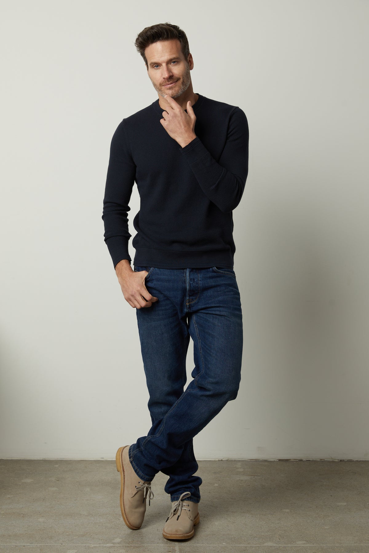   A man is posing in Velvet by Graham & Spencer jeans and a WALTER CREW NECK SWEATER. 