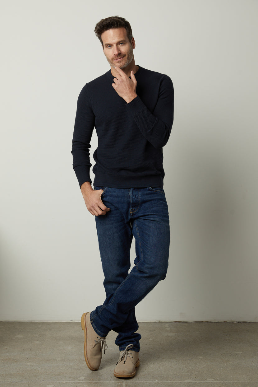 A man stands against a plain background, wearing the Velvet by Graham & Spencer WALTER SWEATER in navy, blue jeans, and brown shoes, with his left hand in his pocket and his right hand touching his chin.