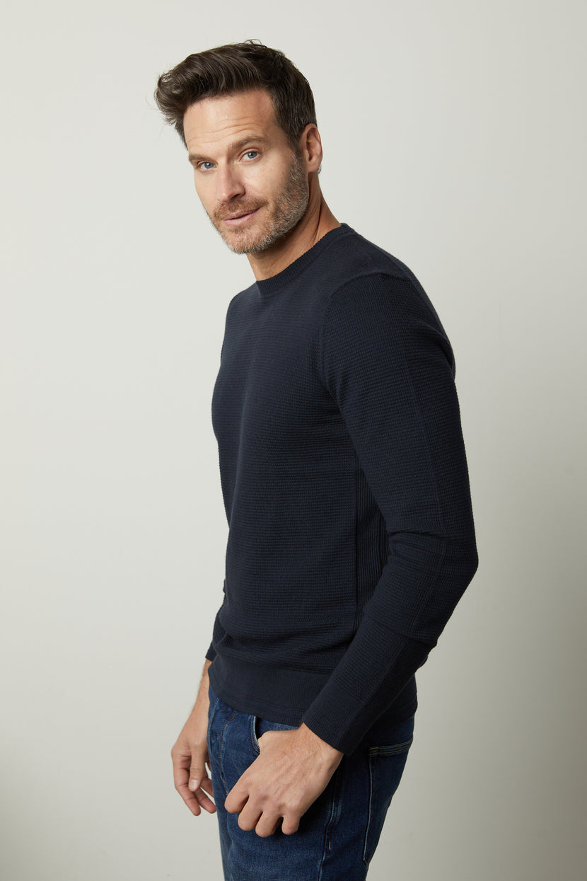 Man wearing a dark blue WALTER SWEATER by Velvet by Graham & Spencer and jeans, standing sideways, looking at the camera.