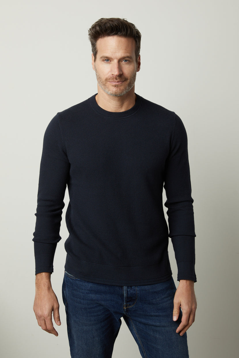 A man with short hair and a beard is wearing the black WALTER SWEATER by Velvet by Graham & Spencer and blue jeans, standing against a plain light-colored background.