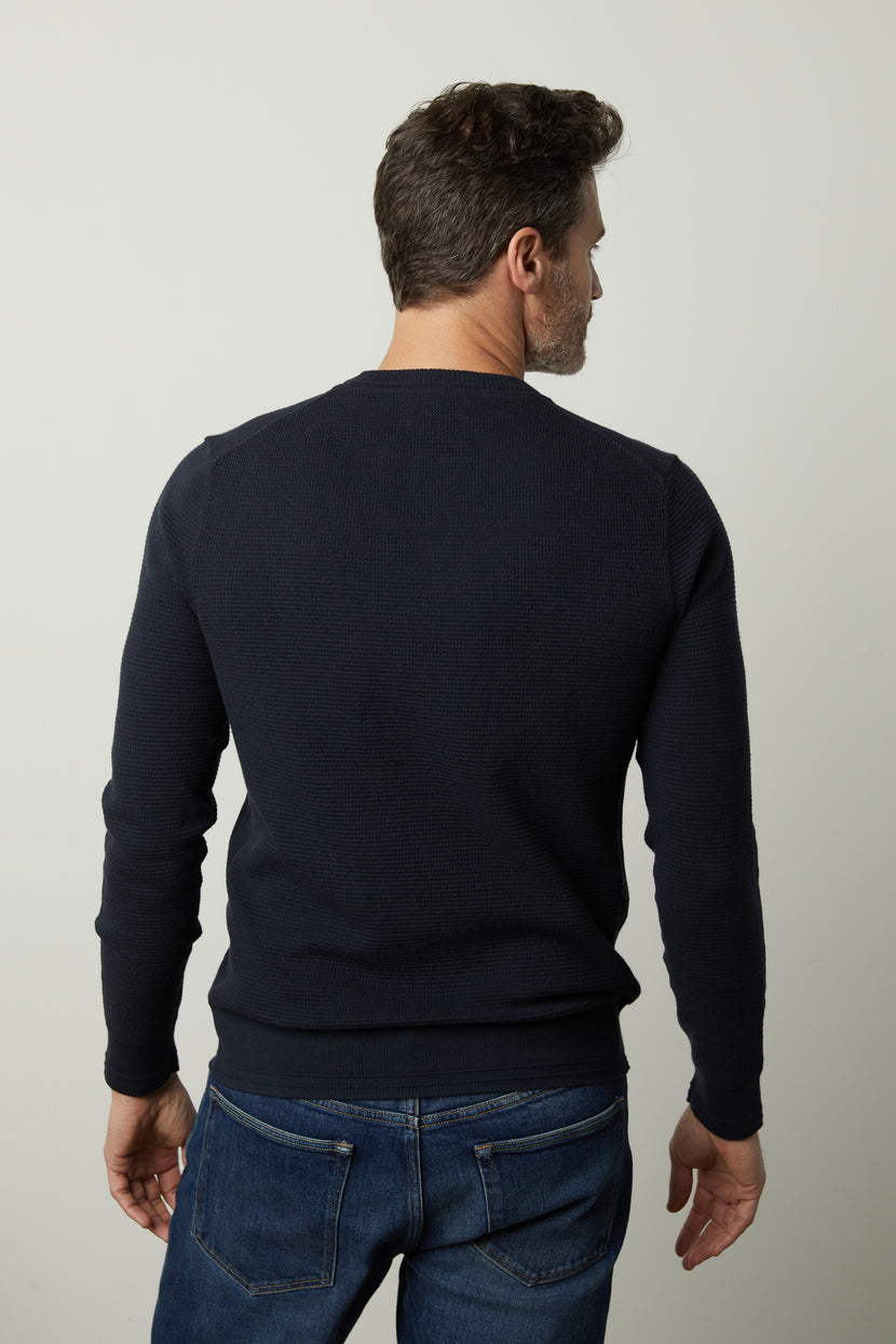 A man with short dark hair, wearing the WALTER SWEATER by Velvet by Graham & Spencer and blue jeans, stands with his back to the camera against a plain background.