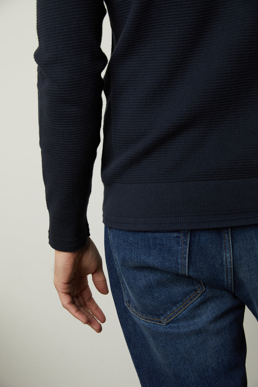 A person wearing the WALTER Sweater by Velvet by Graham & Spencer, a dark blue textured cashmere piece, along with blue jeans is shown from the back with one arm extended downward.